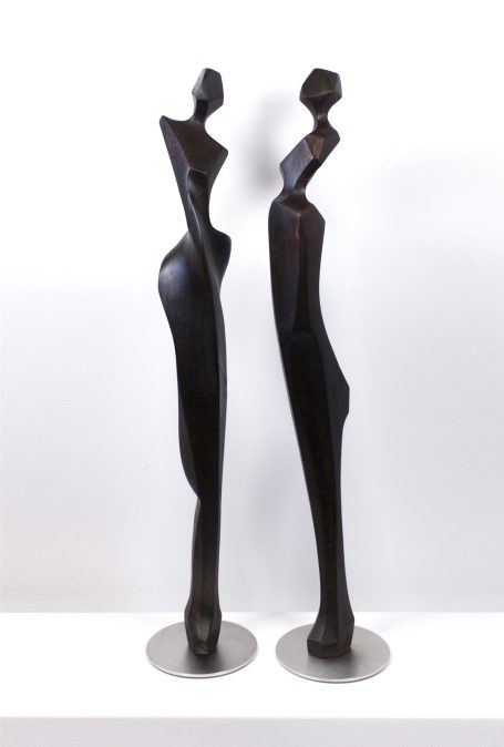 Zwei stilisierte, schlanke Holzstatuen mit eleganten Formen auf Metallständern.